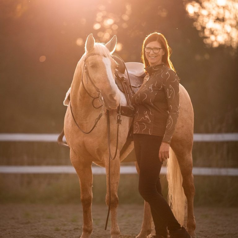 Frau steht neben einem Pferd in der Abenddämmerung, sanftes Licht im Hintergrund.